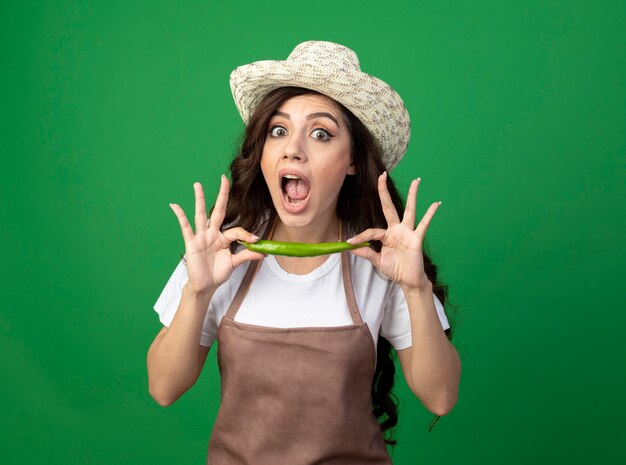 Шокированная молодая женщина-садовник в униформе в садовой шляпе держит острый перец на зеленой стене