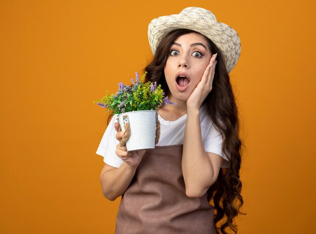 ガーデニング帽子をかぶって制服を着たショックを受けた若い女性の庭師は、顔に手を置き、コピースペースでオレンジ色の壁に隔離された植木鉢を保持します。