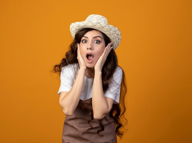 Shocked young female gardener in uniform wearing gardening hat puts hands on face isolated on orange wall with copy space