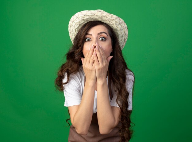 Shocked young female gardener in uniform wearing gardening hat puts hands on mouth isolated on green wall with copy space