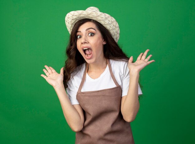 Shocked young female gardener in uniform wearing gardening hat stands with raised hands isolated on green wall with copy space