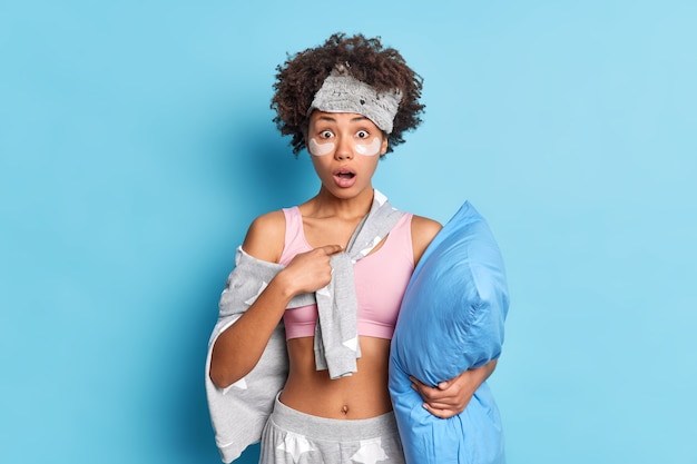 Shocked young female indicates at herself with fore finger surprised to be accused of fault keeps jaw dropped dressed in slumber suit holds pillow isolated over blue wall
