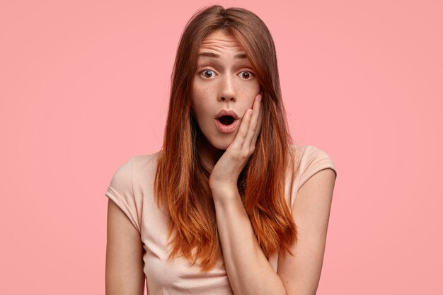 Shocked young female with freckled looks nervously and confusingly at camera