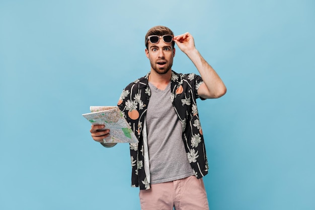 Free photo shocked young guy in modern tshirt and pineapple and palm print shirt looking into camera and holding map on blue backdrop