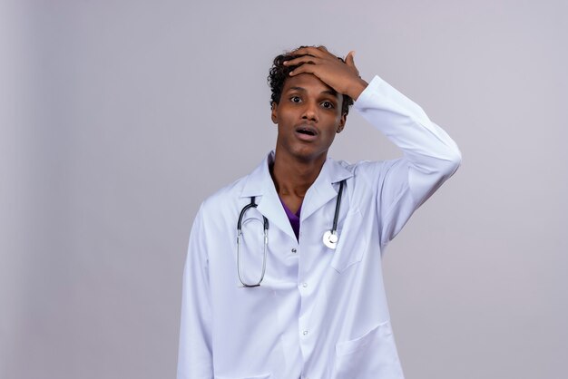 A shocked young handsome dark-skinned doctor with curly hair wearing white coat with stethoscope keeping palm on forehead 