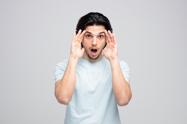Shocked young handsome man looking at camera touching face with both hands isolated on white background