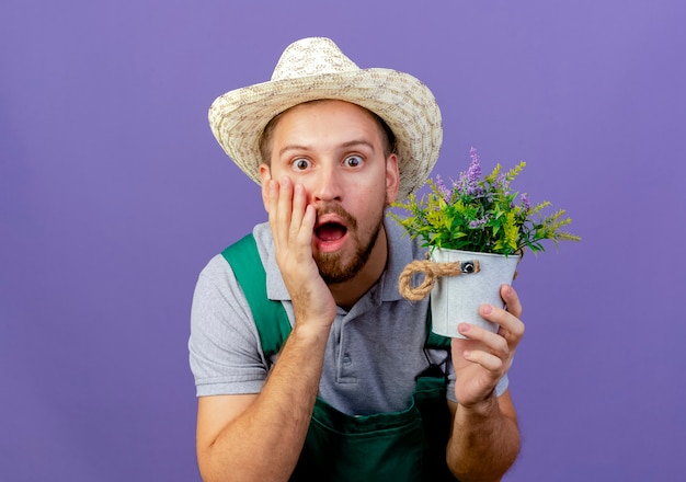 Shocked young handsome slavic gardener in uniform and hat holding flowerpot  touching face isolated on purple wall with copy space