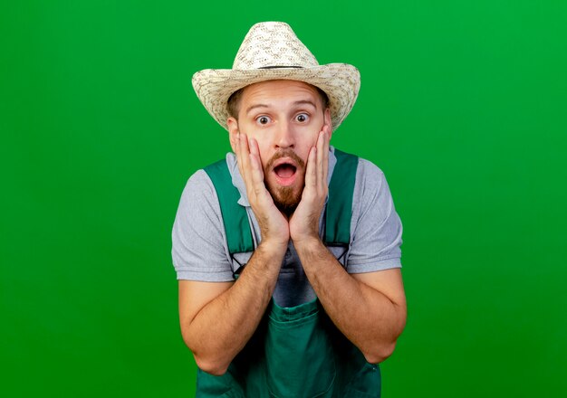 Shocked young handsome slavic gardener in uniform and hat looking  keeping hands on face isolated on green wall with copy space