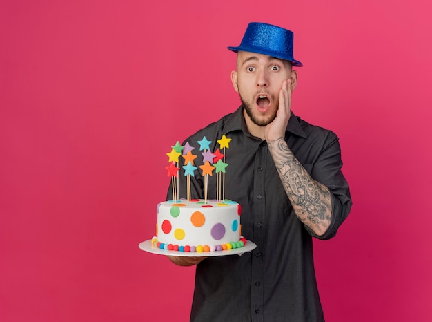 Shocked young handsome slavic party guy wearing party hat holding birthday cake with stars keeping hand on face looking at camera isolated on crimson background with copy space