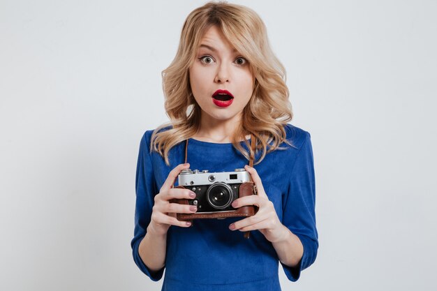 Shocked young lady holding camera over white wall