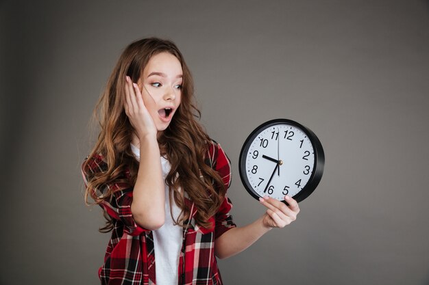 Shocked young lady holding clock. Looking aside.