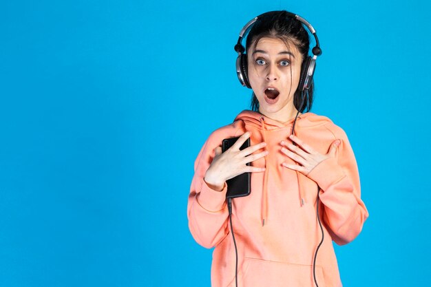 Shocked young lady put her hands to her chest and listening to music on blue background High quality photo