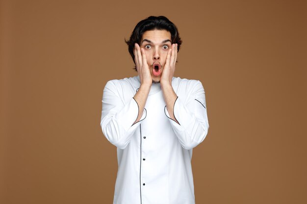 shocked young male chef wearing uniform keeping hands on face looking at camera isolated on brown background