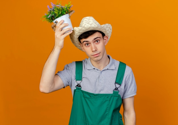ガーデニング帽子をかぶってショックを受けた若い男性の庭師は頭上に植木鉢を保持します