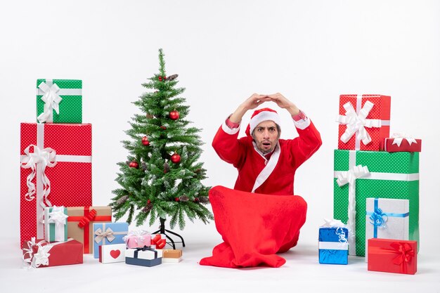 Шокированный молодой человек празднует новый год или рождество, сидя на земле возле подарков и украшенной рождественской елки