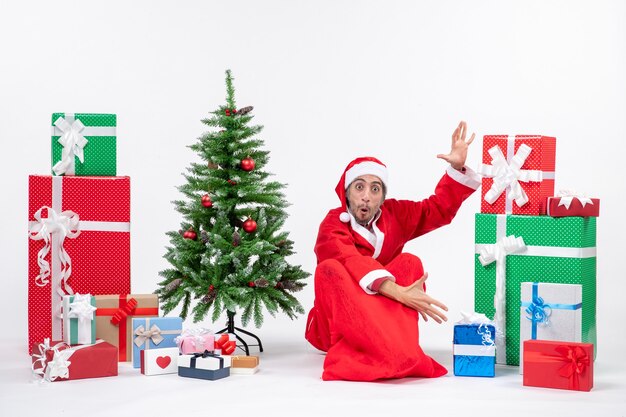 Shocked young man dressed as Santa claus with gifts and decorated Christmas tree sitting on the ground on white background