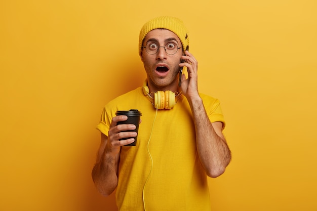 Shocked young man has phone conversation, gasps nervously, stares with panic, wears casual t shirt, hat and spectacles