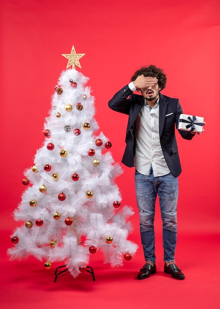 Shocked young man holding his gift closing his eye standing near decorated white Xmas tree on the right side of red