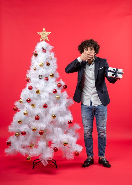 Shocked young man holding his gift and closing his mouthstanding near decorated white Xmas tree on the right side of red