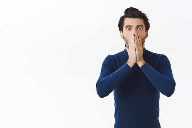 Shocked young man losing lots money, failed business meeting, hold hands on mouth, stare speechless and concerned, gasping worried, nervously react to terrible situation, white wall