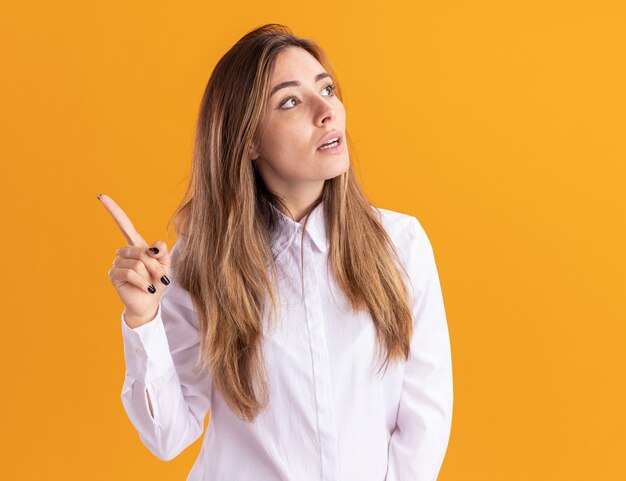 Shocked young pretty caucasian girl looks at side and points up isolated on orange wall with copy space