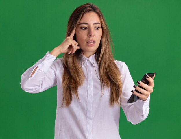 Shocked young pretty caucasian girl puts finger on temple holding and looking at phone on green 