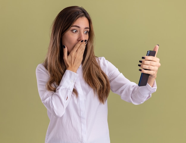 Shocked young pretty caucasian girl puts hand on mouth holding and looking at phone