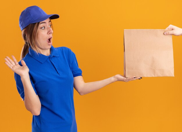 Shocked young pretty delivery girl in uniform takes paper package from someone on orange