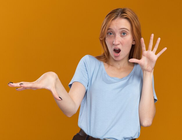 Shocked young redhead ginger girl with freckles holding hands open isolated on orange wall with copy space