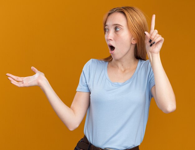 Shocked young redhead ginger girl with freckles holds hand open and points up looking at side isolated on orange wall with copy space