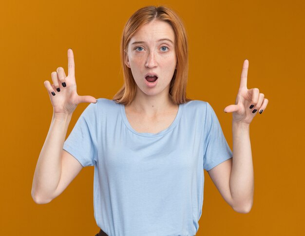 Shocked young redhead ginger girl with freckles pointing up with two hands