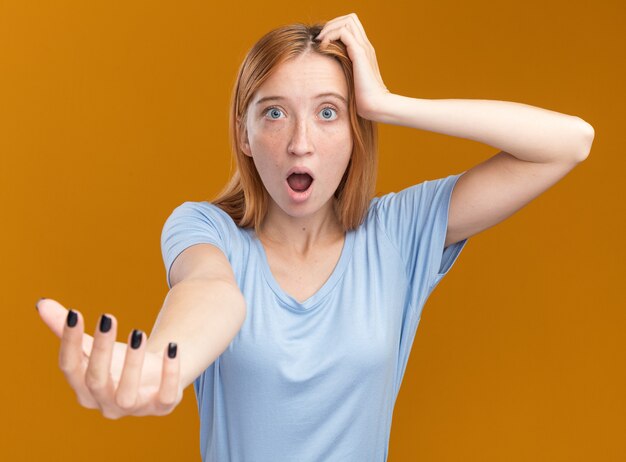 Shocked young redhead ginger girl with freckles puts hand on head and holds hand open