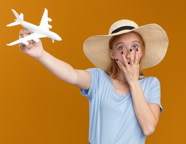 Shocked young redhead ginger girl with freckles wearing beach hat puts hand on mouth and holds model plane