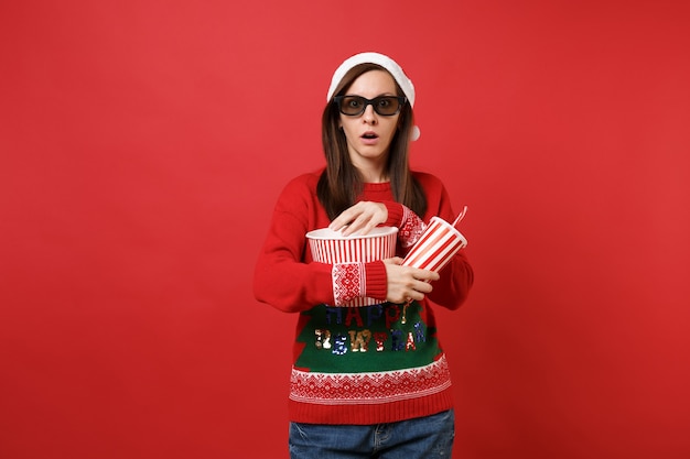 Shocked young santa girl in 3d imax glasses watching movie film, holding popcorn cup of soda isolated on red wall background. happy new year 2019 celebration holiday party concept. mock up copy space.