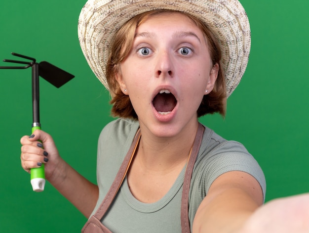 Foto gratuita giovane giardiniere femminile slavo scioccato che indossa cappello da giardinaggio che tiene il rastrello della zappa e che prende selfie isolato sulla parete verde con lo spazio della copia