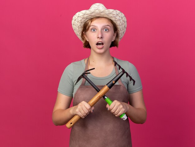 Shocked young slavic female gardener wearing gardening hat holding rake and hoe rake