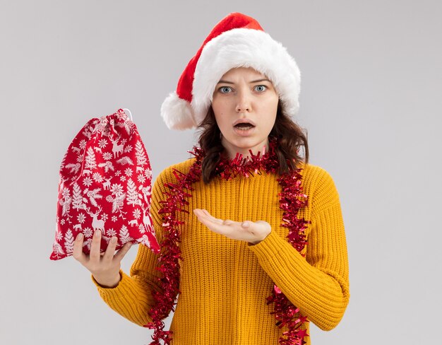 Shocked young slavic girl with santa hat and with garland around neck holding christmas gift bag and keeping hand open isolated on white background with copy space