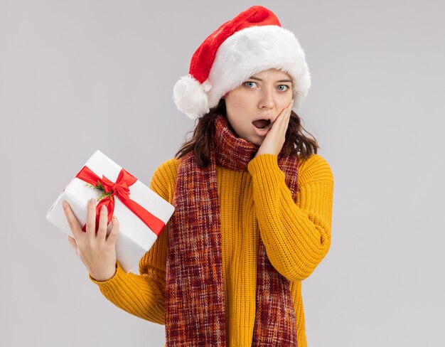 산타 모자와 목에 스카프로 충격을받은 젊은 슬라브 소녀는 얼굴에 손을 넣고 복사 공간이 흰 벽에 고립 된 크리스마스 선물 상자를 보유하고 있습니다.