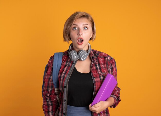 Shocked young slavic student girl with headphones wearing backpack holds book and notebook 