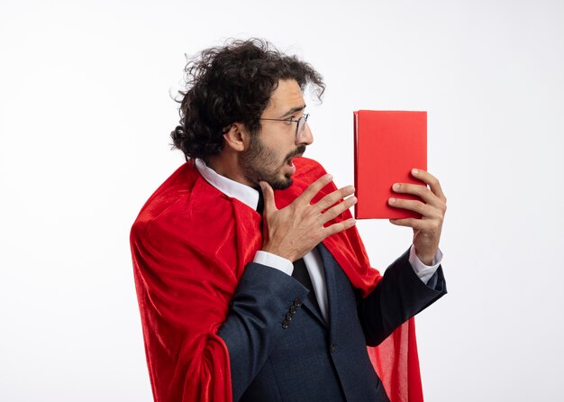 Shocked young superhero man in optical glasses wearing suit with red cloak looks and points at book isolated on white wall