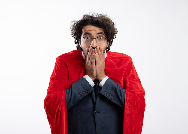 Free photo shocked young superhero man in optical glasses wearing suit with red cloak puts hands on mouth isolated on white wall