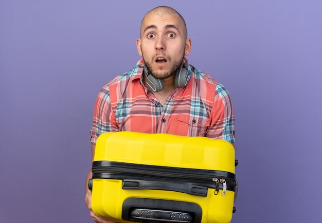 shocked young traveler man with headphones around his neck holding suitcase isolated on purple wall with copy space