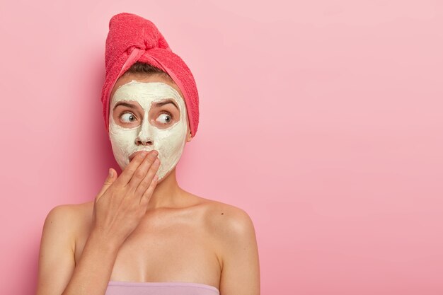 Shocked young woman applies clay nourishing facial mask, covers mouth with palm, hydrates and calms skin, wears rosy towel wrapped on head, stands against pink wall. Rejuvenation concept