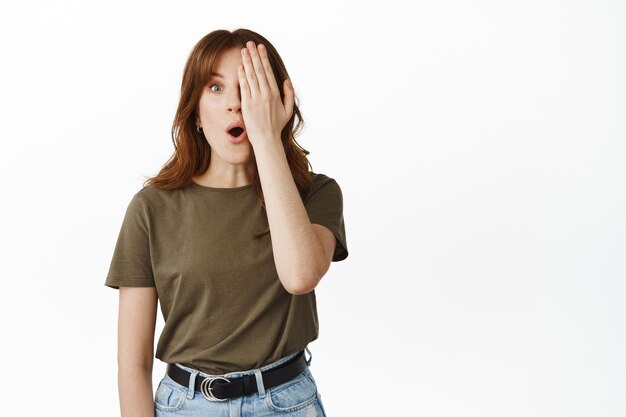 Shocked young woman con half of face with hand, drop jaw and gasp, checking out something amazing, standing on white