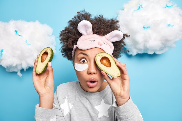 Shocked young woman covers eyes with halves of avocado uses organic product and natural cosmetics applies patches under eyes wears pajama sleepmask poses against blue wall