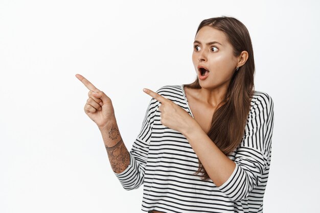 Shocked young woman drop jaw while looking and pointing fingers left, staartled by something, standing on white.