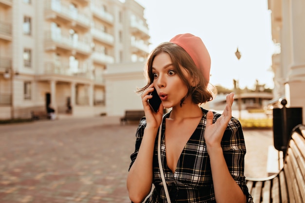 電話で話しているエレガントな衣装でショックを受けた若い女性。通りに立っている魅力的なフランスの女性モデルの屋外の肖像画。