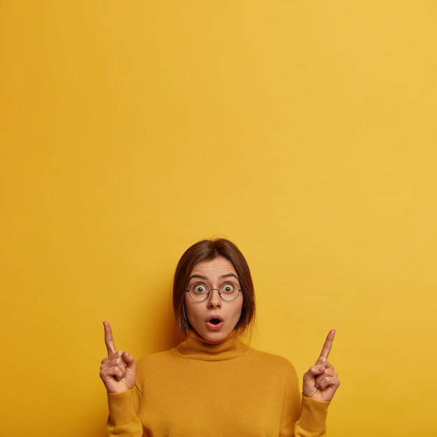 Shocked young woman gossips about latest news, indicates both index fingers upwards, hears surprising news, opens mouth, wears big round glasses and turtleneck, isolated on yellow wall