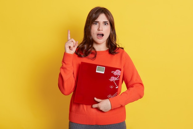 Shocked young woman holding scale and pointing up with index finger, having shocked facial expression, does not like her weight, being shocked and upset.