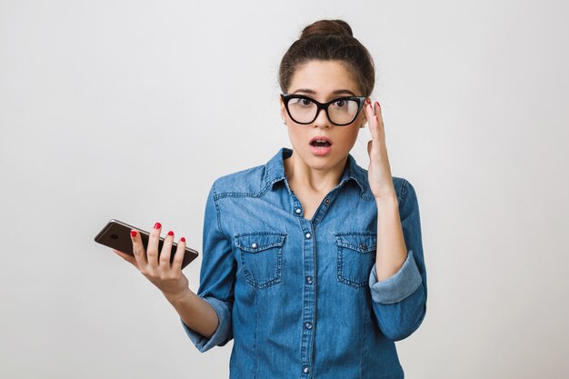 Shocked young woman holding smart phone, wearing stylish glasses, opened mouth, , surprised face expression, isolated , denim shirt